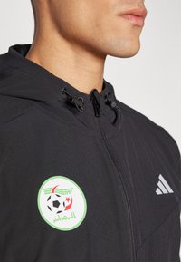 Crni sportski sako s kapuljačom, s okruglim zeleno-belim nogometnim emblemom Alžira i detaljem sivi Adidas loga na ramenu.