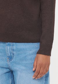 Brun strikket genser med ribbestrikkede mansjetter, parret med lyseblå jeans. Nærbilde av erm og hånd viser teksturert stoff.
