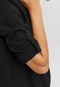 Chemise en lin noir avec manches retroussées, dotée d'une fermeture à boutons sur le poignet. Texture lisse avec une coupe décontractée.