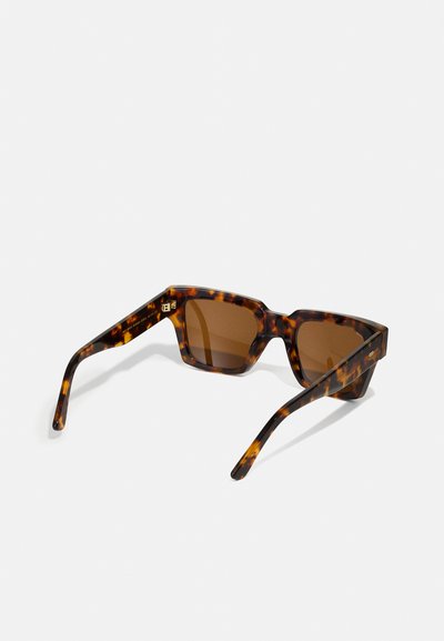 Lunettes de soleil en écaille de tortue avec une monture géométrique épaisse, des verres teintés marron et des branches épurées, présentant un logo subtil sur la branche.