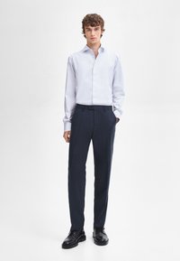 Witte longsleeve shirt met knopen aan de voorkant, gecombineerd met donkerblauwe broek en zwarte nette schoenen, wat een formele outfit creëert.