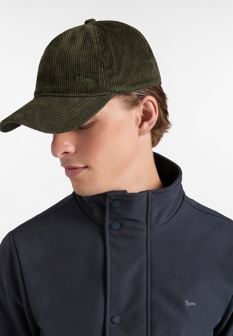 Cappello in corda verde oliva con visiera curva e superficie testurizzata, con un piccolo logo sulla parte anteriore. Indossato con una giacca scura.