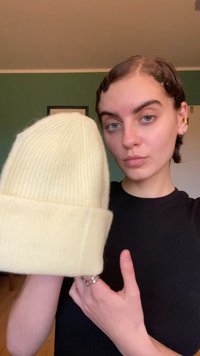 Svijetložuta pletena kapa s rebrastim uzorkom i preklopom, prikazana u ruci nasuprot tamnoj majici, naglašavajući svoju teksturu i oblik.