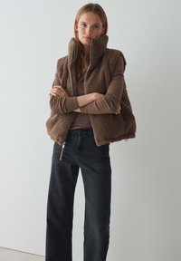 Braune Fleece-Weste mit hohem Kragen, Reißverschluss und seitlichen Taschen, kombiniert mit einem braunen Langarmoberteil und schwarzen weit geschnittenen Jeans.