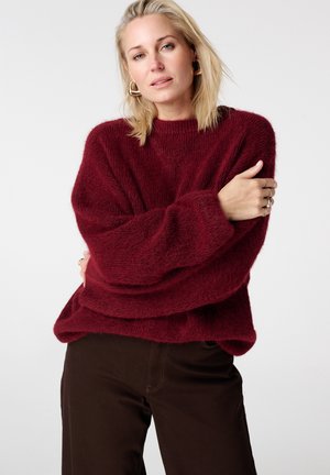 Pull en maille bordeaux avec manches surdimensionnées et tissu texturé, associé à un pantalon marron. Léger, coupe ample. Pas de motifs remarquables.