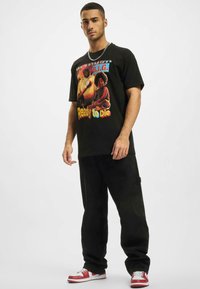 T-shirt en coton noir avec un imprimé graphique de deux figures et le texte "Ready to die" en polices colorées. Associée à un pantalon noir et des baskets rouges.