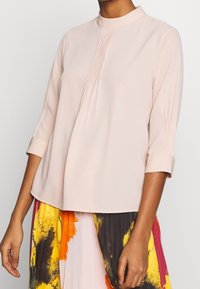 Blouse rose clair avec un col montant et des manches trois-quarts. Présente de subtils plis à l'avant et une texture lisse, complétée par des poignets retroussés.