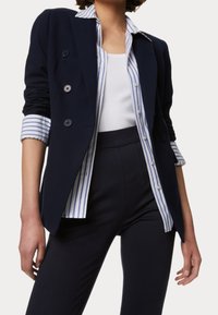 Námořnický blejzr s dvojitými knoflíky, texturovanou tkaninou a kontrastními pruhovanými manžetami, nošený přes bílý top bez rukávů a tmavé kalhoty.