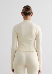 Top fitted color crema a coste con maniche lunghe e bordi arricciati, abbinato a pantaloni coordinati. Pattern testurizzato su tutta la superficie.
