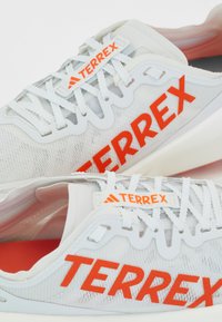 White athletic shoes featuring a mesh upper, orange "TERREX" branding, flat laces, and a cushioned sole with a subtle gradient.