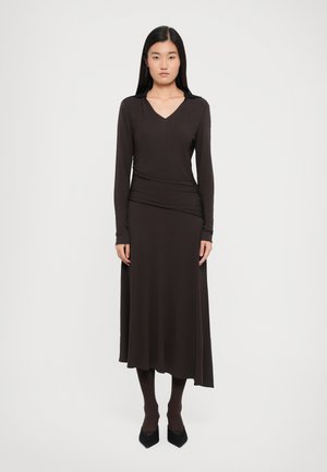 Long-sleeve, V-neck brown dress with a fitted bodice, draped waist, and flowing skirt. Smooth fabric with a soft texture. Black pointed shoes.
