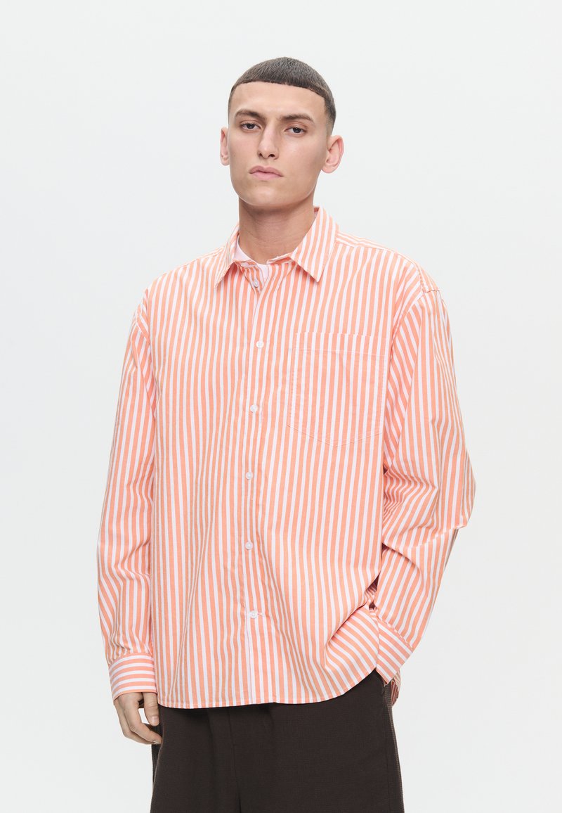 Young man wearing an orange and white vertical striped button-up shirt with one hand in pocket against plain background.