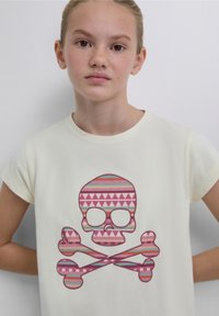 Cremefarbenes T-Shirt mit kurzen Ärmeln, das ein großes, pink-blau gemustertes Totenkopfdesign mit gekreuzten Knochen zeigt, vor einem neutralen Hintergrund.