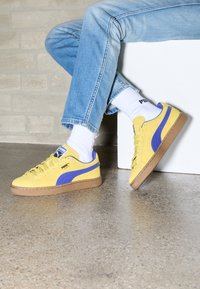 Yellow suede sneakers with purple accents and beige rubber soles, featuring round laces and a Puma logo on the side. Worn with white socks.