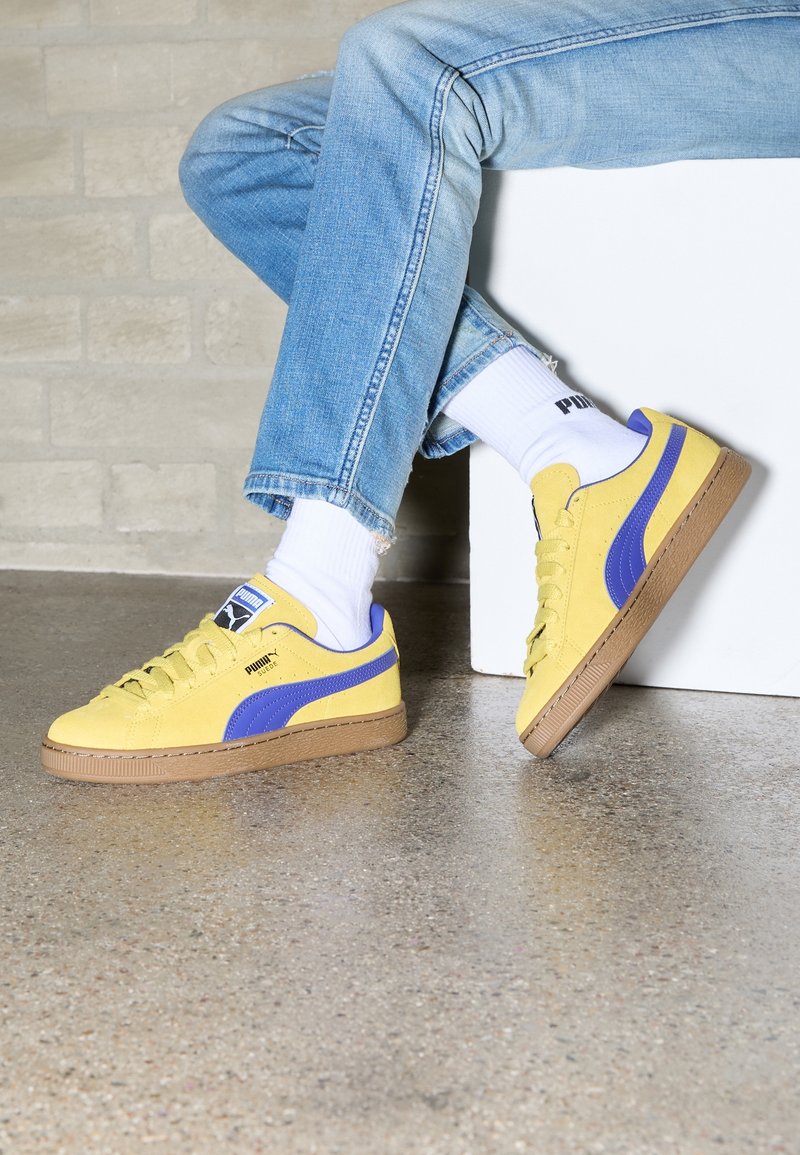 Yellow suede sneakers with purple accents and beige rubber soles, featuring round laces and a Puma logo on the side. Worn with white socks.