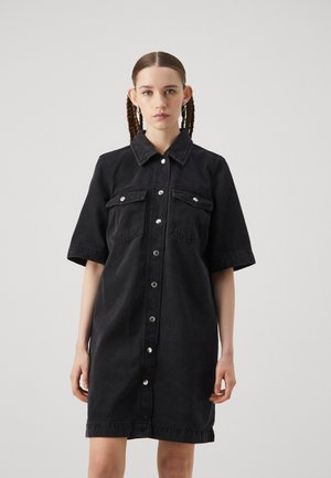 Robe chemise en denim noir à manches courtes, patte de boutonnage, deux poches poitrine. Texture lisse, coupe légèrement décontractée, longueur au genou.