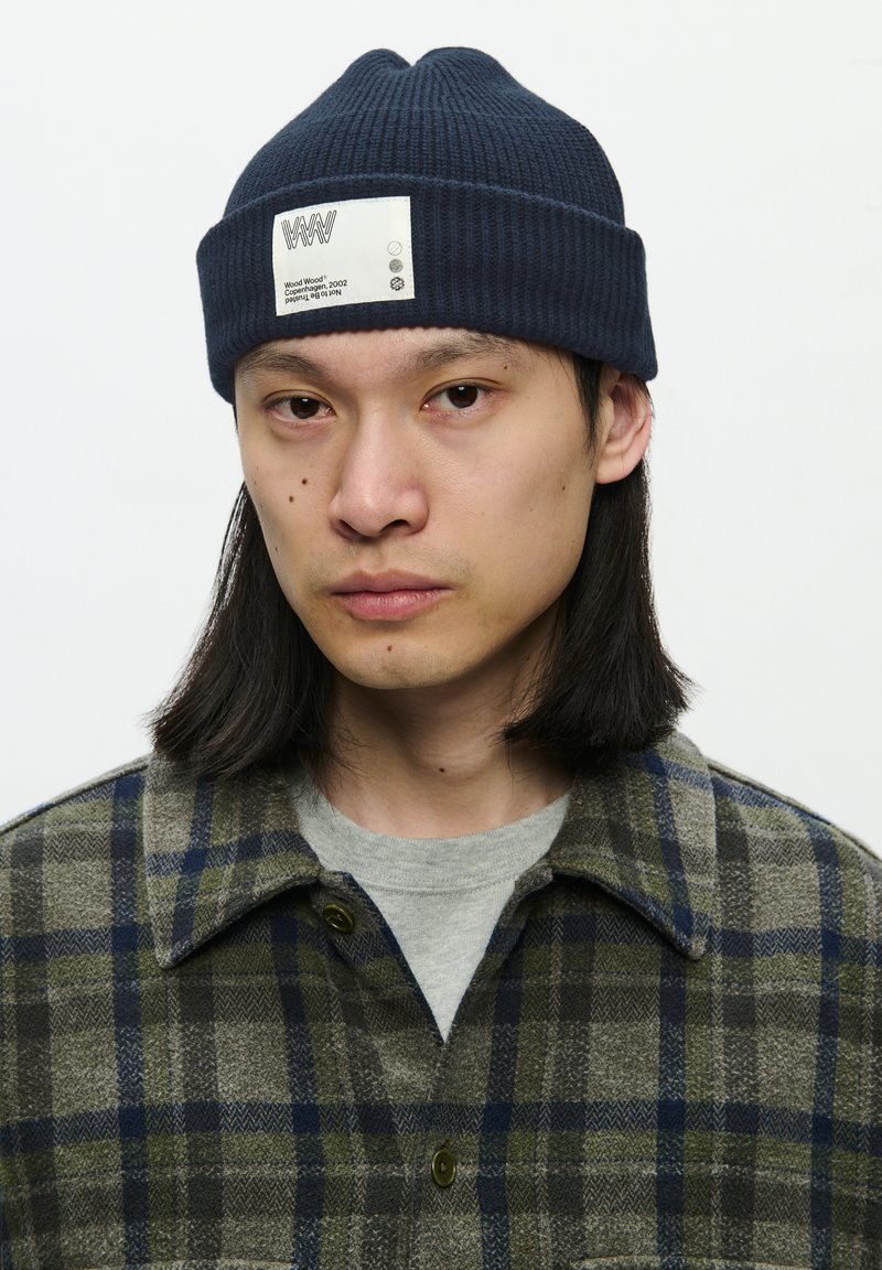Navy ribbed beanie with a white label, worn with a plaid olive-green shirt featuring blue accents and a gray crew neck shirt underneath.
