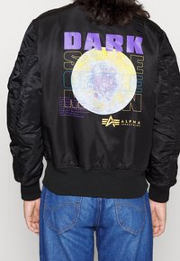 Czarna kurtka bomber z prążkowanymi mankietami, z dużym nadrukiem księżyca i napisem "DARK SIDE OF THE MOON" w kolorze fioletowym i żółtym na plecach.