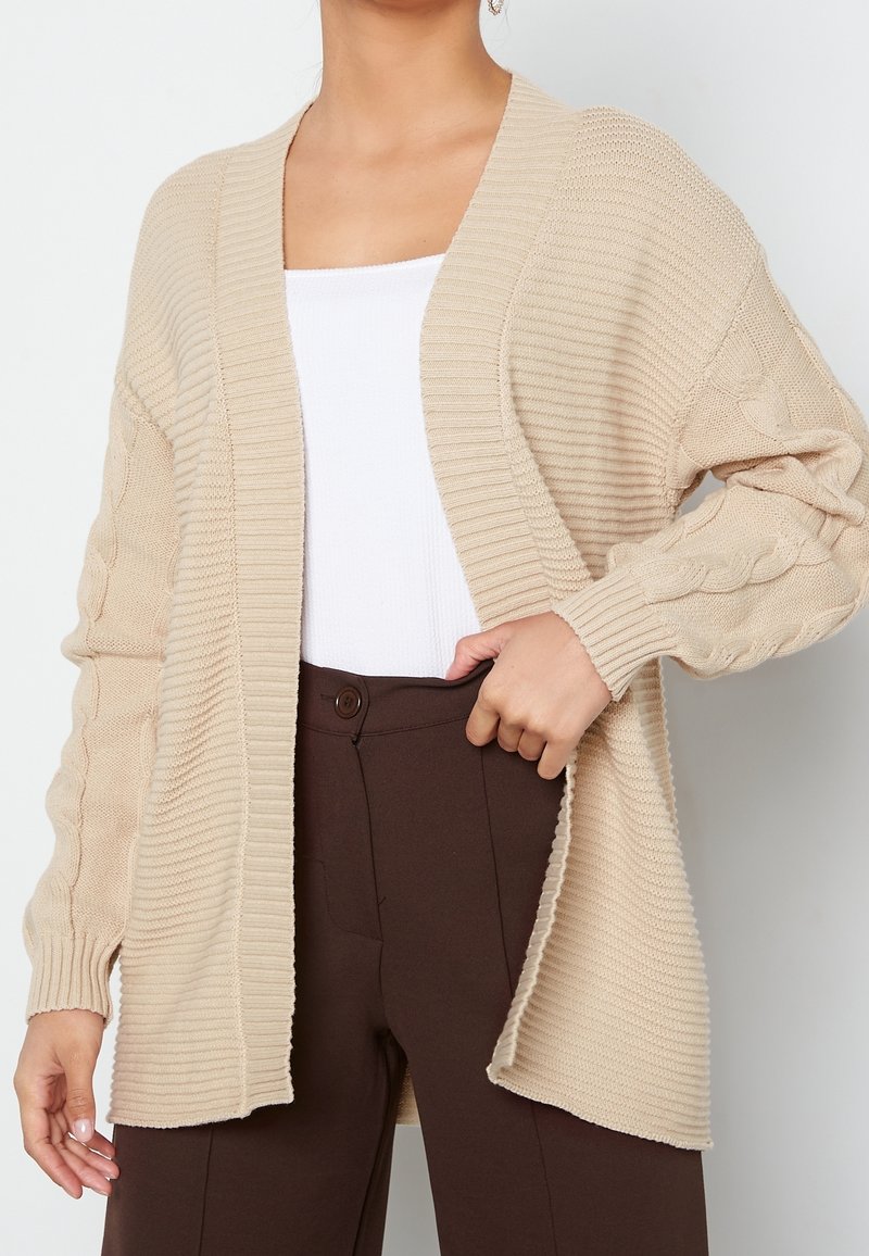 Cardigan beige en maille ouvert à l'avant avec des motifs torsadés sur les manches, porté par-dessus un haut blanc et un pantalon taille haute marron foncé.