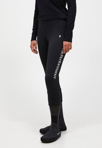 Zwarte leggings hebben een nauwsluitende pasvorm, een hoge tailleband en een wit logo "Peak Performance" aan de linkerkant. Geverfde sokken en zwarte schoenen maken de uitstraling compleet.