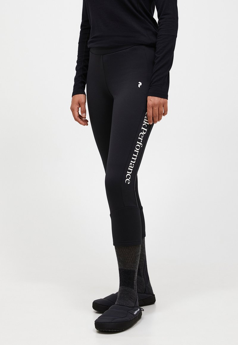 Zwarte leggings hebben een nauwsluitende pasvorm, een hoge tailleband en een wit logo "Peak Performance" aan de linkerkant. Geverfde sokken en zwarte schoenen maken de uitstraling compleet.
