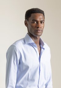 Camisa abotonada de color azul claro con un patrón de cuadros finos, cuello estructurado y mangas largas. La tela parece suave y ligera.