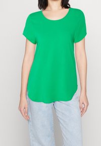Grünes Kurzarmoberteil mit rundem Halsausschnitt aus glattem Stoff; kombiniert mit hellblauen, locker sitzenden Jeans.