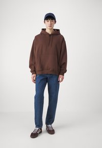 Brun hette med en frontlomme, kombinert med blå denimjeans og mørkerøde sneakers. Modell iført en blå caps, står mot en nøytral bakgrunn.