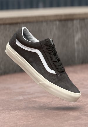 Zwarte suède skate-schoen met witte zijstreep en crèmekleurige rubberen zool, getoond in de lucht tegen een stenen achtergrond.