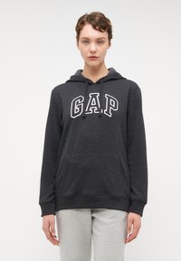 Aktīvās melnas pelēkās krāsas kapuča krekls ar baltu "GAP" uzrakstu, kangoru kabatu, aukliņu kapuci un ribotu piedurkņu apmali. Valkāts kopā ar gaiši pelēkiem sporta biksēm.