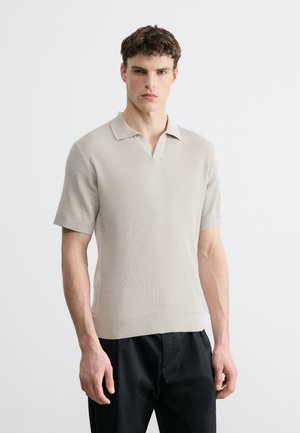 Młody mężczyzna ubrany w beżową, krótko rękawową dzianinową koszulkę polo i czarne spodnie, stojący na jednolitym białym tle.