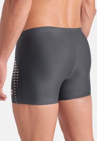 Costumi da bagno da uomo, tessuto grigio scuro, vestibilità aderente, superficie liscia, motivo laterale con accenti triangolari bianchi, logo del marchio sulla gamba sinistra.