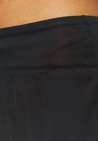 Musta urheilukangas, jossa on sileä pinta, sisältää kokoillan vyötärönauhan ja tyylikkään muotoilun. Lähellä otettu kuva keskittyy kankaan yksityiskohtiin.