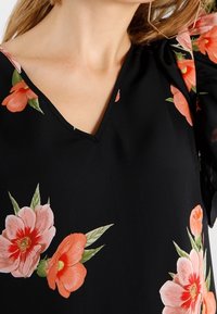 Černá květinová halenka s výstřihem do V a nabíranými rukávy, zdobená velkými růžovými a korálovými květy a zelenými listy.