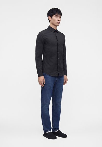 Mann, der leicht nach rechts schaut, trägt ein schwarzes, langärmliges Hemd mit Knöpfen, blaue Jeans, weiße Socken und schwarze Slipper.