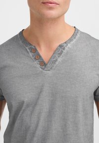 Camiseta de algodón gris con escote en V, que cuenta con tres botones marrones y una textura sutil. Mangas cortas con un corte relajado.