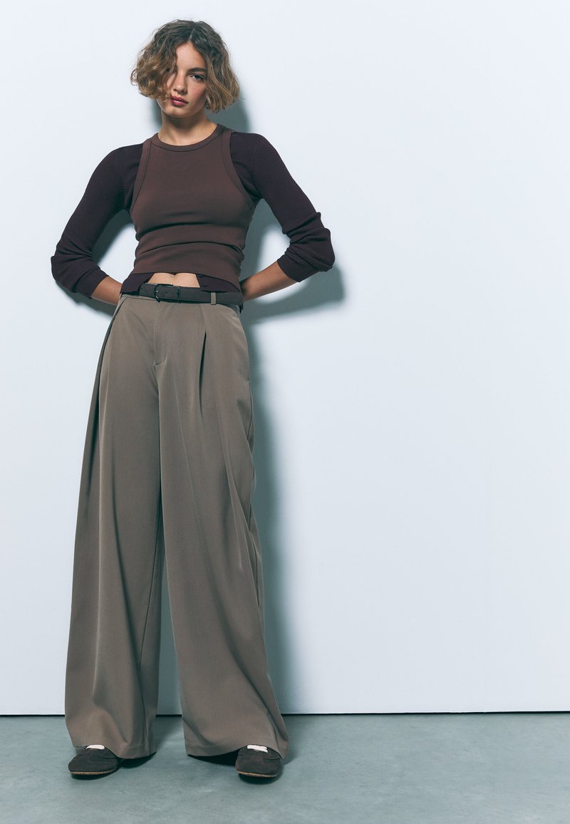 Prilegajoča rjava rebrasta kratka majica, v kombinaciji s širokimi zloženimi bež hlačami. Obleka vsebuje črni pas in temno rjave natikače.