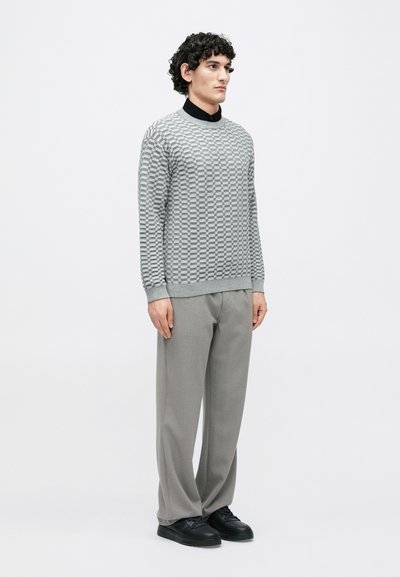 Camisola cinzenta com padrão e gola canelada, combinada com calças cinza claras e ténis pretos. Modelo está em pé contra um fundo simples.