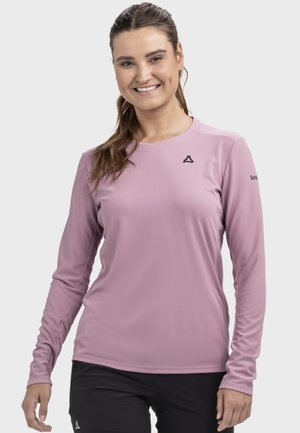 Lächelnde Frau mit braunen Haaren in einem Pferdeschwanz, die ein rosa langärmeliges Sportshirt und schwarze Hosen vor einem einfarbigen Hintergrund trägt.