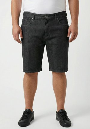 Mand iført sorte denimshorts, hvid t-shirt og sorte sneakers, står med armene afslappet ned langs siden på hvid baggrund.