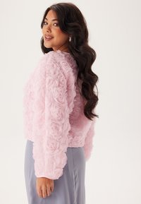 Ljusrosa kort jacka i texturerat tyg med blommig applikation, med långa ärmar och en figurnära design. Bärs över en grå kjol.