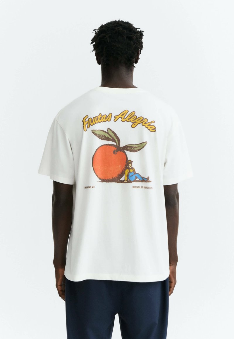 Homme à la peau foncée et aux cheveux courts torsadés, portant un t-shirt blanc avec un gros fruit orange et le texte "Frutas Alegría" dans le dos.