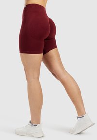 Vrouw in strakke bordeauxrode sportshorts en witte sneakers, staand met een been naar achteren gestrekt tegen een effen achtergrond.