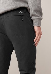 Pantalon d'extérieur noir en tissu résistant, doté d'une coupe ajustée, de poches zippées et d'une taille soulignée par un logo.