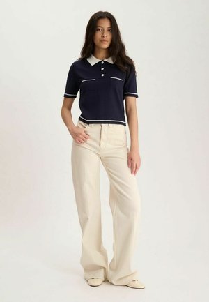Femme debout vêtue d'un polo bleu marine à manches courtes avec bordure blanche et d’un pantalon large crème sur un fond clair uni.