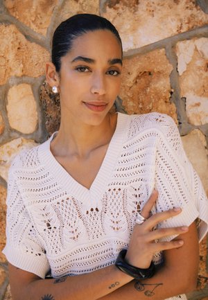 Tumejuustega naine valges mustrilises kootud topis seisab kivimüüri taustal, kandmas hõbedaid kõrvarõngaid, sõrmuseid ja musta käevõru, nähtavate tätoveeringutega.