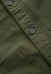 Gros plan sur un tissu vert olive avec deux boutons gris foncé et une poche cousue sur un vêtement.