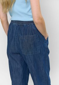 Pantaloni din denim cu un brâu elastic, de culoare albastră, având un singur buzunar pe spate și o croială lejeră. Țesătura are o ușoară textură.