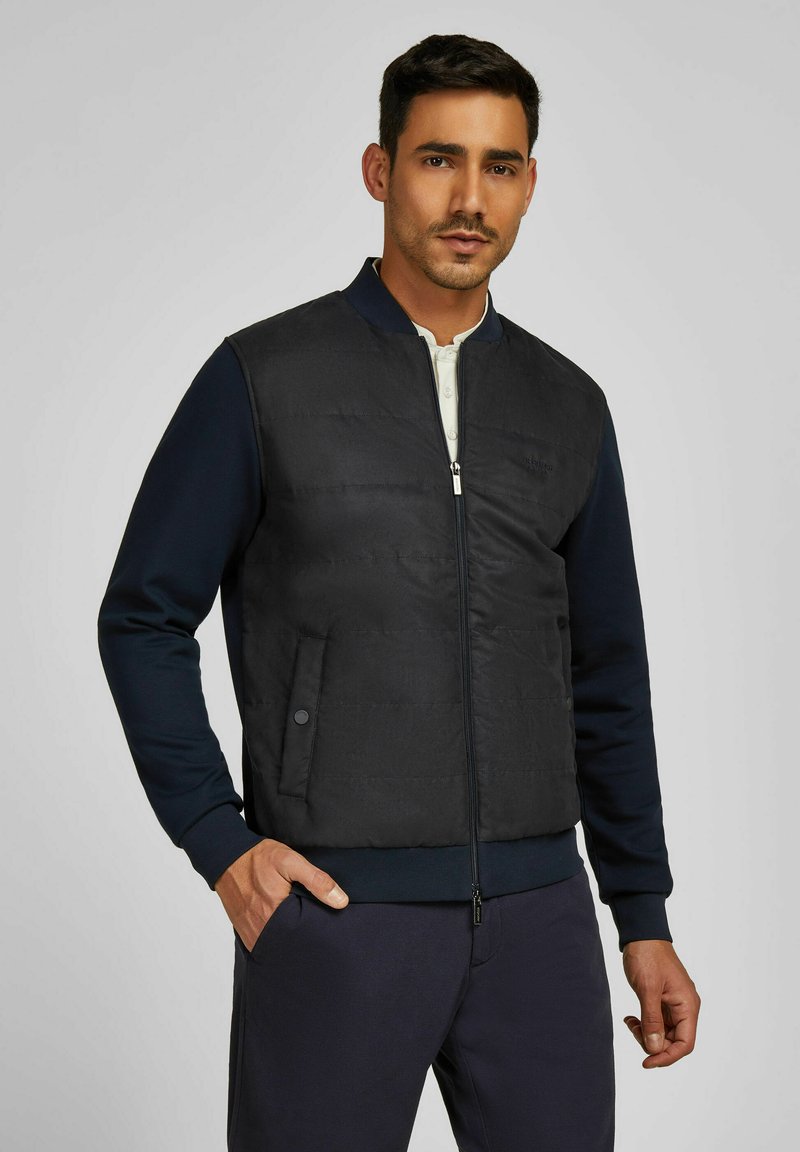 Schwarze gesteppte Bomberjacke mit navyblauem Rippkragen und -bündchen, ausgestattet mit einem Reißverschluss auf der Vorderseite und seitlichen Taschen.