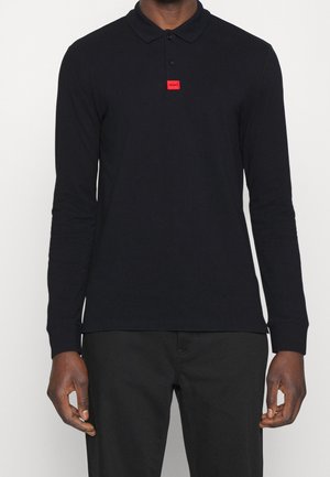 Mand iført en sort langærmet polo med et lille rødt "HUGO" logo på brystet og sorte bukser mod en hvid baggrund.